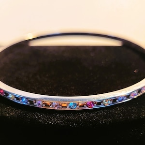 May include: A silver-toned bangle bracelet with a row of colorful gemstones. The gemstones are a mix of pink, blue, yellow, and purple. The bracelet is set against a dark background.