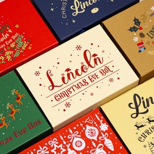 Könnte beinhalten: Eine Sammlung rechteckiger Geschenkboxen in verschiedenen Farben wie Rot, Marineblau, Kraftbraun, Creme und Grün. Jede Box ist mit festlichen Weihnachtsmotiven wie Rentieren, Ornamenten und Schneeflocken verziert, wobei der Name Lincoln und Christmas Eve Box in dekorativer Schrift und Blockbuchstaben aufgedruckt sind.