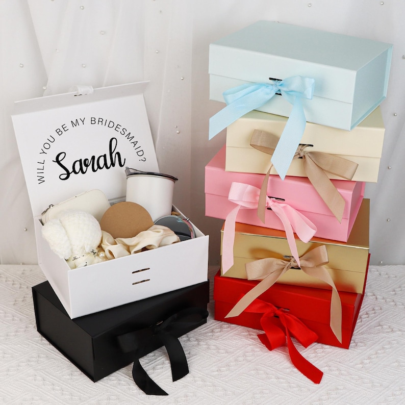 May include: A stack of gift boxes in various colors, including white, black, light blue, pink, and gold. Each box is tied with a matching ribbon. The top box is open, revealing gifts and the text "Will you be my Bridesmaid? Sarah".