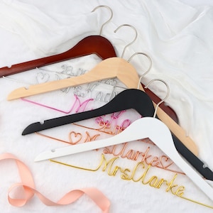 May include: Four wooden hangers with wire lettering that says "Mrs. Clarke" on the white hanger, "Mrs. Mack" on the natural wood hanger, and "Mrs." on the black hanger. The hangers are arranged in a stack with the white hanger on top.