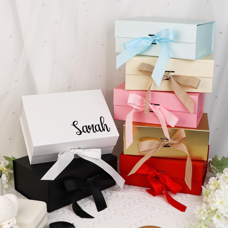 May include: A collection of gift boxes in various colors, including white, black, gold, pink, red, and light blue. Each box is adorned with a matching satin ribbon bow. The word "Sarah" is written on the white box.