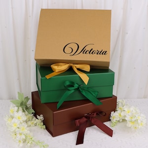 May include: Three stacked gift boxes in brown, green, and maroon, each tied with a ribbon. The top box is brown with the name "Victoria" in black script. The green box has a gold ribbon, the middle box has a green ribbon, and the bottom box has a maroon ribbon.