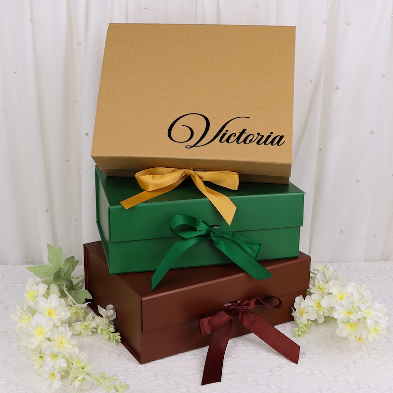 May include: Three stacked gift boxes in brown, green, and maroon, each tied with a ribbon. The top box is brown with the name "Victoria" in black script. The green box has a gold ribbon, the middle box has a green ribbon, and the bottom box has a maroon ribbon.