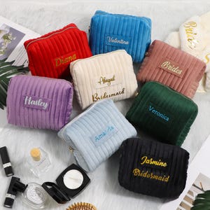 May include: A collection of corduroy cosmetic bags in various colors, including red, blue, purple, cream, pink, green, light blue, and black. Each bag is rectangular with a zipper closure and features a name embroidered in a contrasting color. Some bags have the word "Bridesmaid" embroidered.
