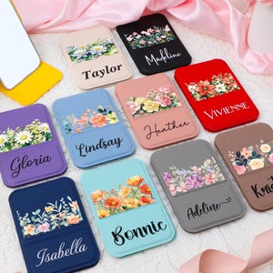 May include: A collection of floral phone card holders in various colors, including purple, blue, and pink. Each holder features a different name in a script font, such as "Taylor" and "Vivienne", and a floral design.