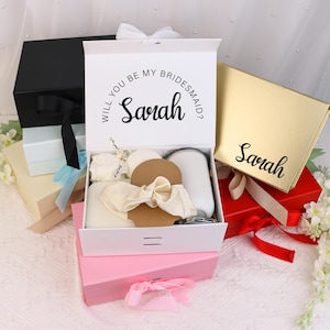 May include: A white gift box with the text "Will you be my bridesmaid? Sarah" is open, revealing various items. Other boxes are visible, including black, gold, and pink, some with ribbons. The theme suggests a bridal party gift set.