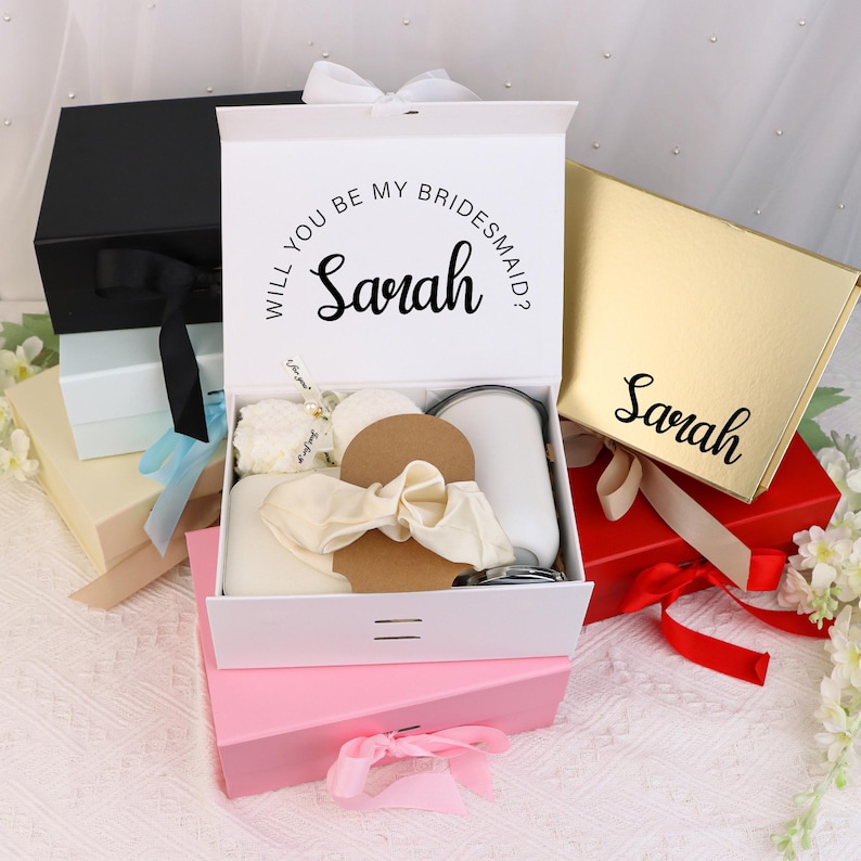 May include: A white gift box with the text "Will you be my bridesmaid? Sarah" is open, revealing various items. Other boxes are visible, including black, gold, and pink, some with ribbons. The theme suggests a bridal party gift set.