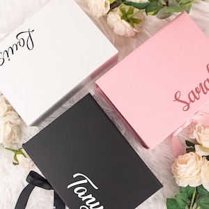 May include: Three gift boxes in white, pink, and black, each with a name in cursive script. The white box has the name "Louis", the pink box has "Sarah", and the black box has "Tany". The boxes are on a white, fluffy surface with floral accents.