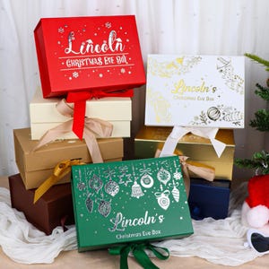 May include: A collection of rectangular gift boxes in red, cream, brown, white, gold, and green. Each box features a ribbon closure and personalized text reading Lincoln's Christmas Eve Box, decorated with silver or gold foil holiday motifs.