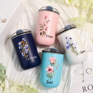 Könnte beinhalten: Vier personalisierte isolierte Becher in Marineblau, Rosa, Weiß und Türkis. Jeder Becher hat ein Blumendesign und einen Namen: Diane, Juliet und Natalia. Die Becher haben einen schwarzen Deckel und sind für heiße oder kalte Getränke konzipiert.