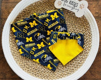 May include: A collection of dog bandanas and a bone-shaped sign. The bandanas are navy blue with a yellow and white pattern of the word "MICHIGAN", the letter "M", and football helmets. The sign says "DOG KISSES fix everything".