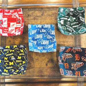 May include: Five fabric items with sports team logos. The items include Detroit Red Wings, Detroit Lions, Michigan State Spartans, Michigan Wolverines, and Detroit Tigers. The items are in red, blue, green, and black.