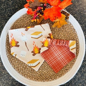 May include: Two triangular pet bandanas featuring a cream-colored fabric with a repeating pattern of cartoon campfires, marshmallows, and s'mores, and a second bandana with a rust-orange and white plaid pattern and a gathered elastic collar.