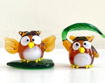 May include: Two small glass owl figurines with brown and white bodies, yellow beaks, and large white eyes. One owl is holding a green leaf over its head. Both owls are sitting on green leaf bases.