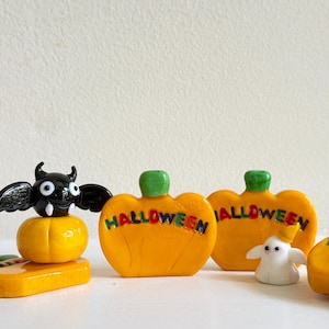 May include: A collection of glass figurines including a black bat with white fangs perched on a yellow pumpkin, two orange pumpkins with green stems and the word HALLOWEEN in colorful letters, and a small white ghost wearing a yellow hat.