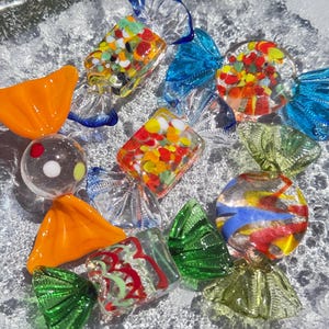 May include: A collection of colorful glass candy pieces in various shapes, including wrapped candies and round lollipops. The candies feature vibrant colors like red, yellow, blue, and green, and are displayed on a textured, clear surface.