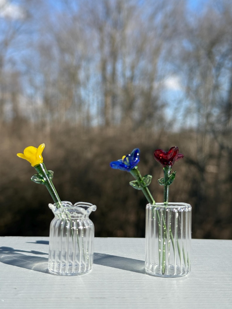 K&ouml;nnte beinhalten: Drei Glasvasen, jede mit einer einzelnen Glasblume. Die Blumen sind gelb, blau und rot. Die Vasen sind aus klarem Glas mit einer gerippten Textur.