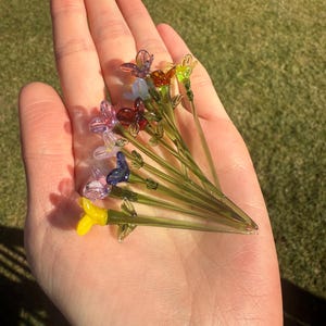 K&ouml;nnte beinhalten: Eine Sammlung von zehn Glasblumen-Steckern in verschiedenen Farben, darunter Lila, Blau, Gelb, Gr&uuml;n und Orange. Die Blumen sind handgefertigt und haben einen zarten, verspielten Look.