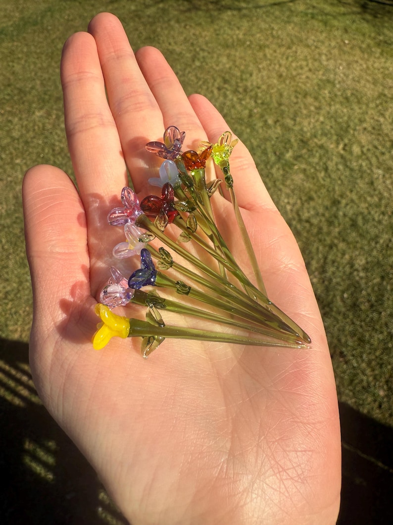 K&ouml;nnte beinhalten: Eine Sammlung von zehn Glasblumen-Steckern in verschiedenen Farben, darunter Lila, Blau, Gelb, Gr&uuml;n und Orange. Die Blumen sind handgefertigt und haben einen zarten, verspielten Look.