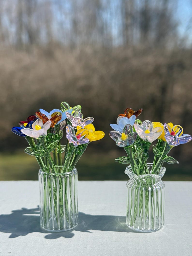 K&ouml;nnte beinhalten: Zwei Glasvasen mit bunten Glasblumenarrangements. Die Vasen sind aus klarem Glas mit vertikalen Linien. Die Blumen sind in Blaut&ouml;nen, Gelb, Rosa und Lila.