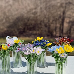 K&ouml;nnte beinhalten: Eine Sammlung bunter Glasblumenstr&auml;u&szlig;e in klaren Glasvasen mit gerippter Textur. Die Blumen sind in verschiedenen Farben angeordnet, darunter Gelb, Rosa, Blau, Lila und Rot.
