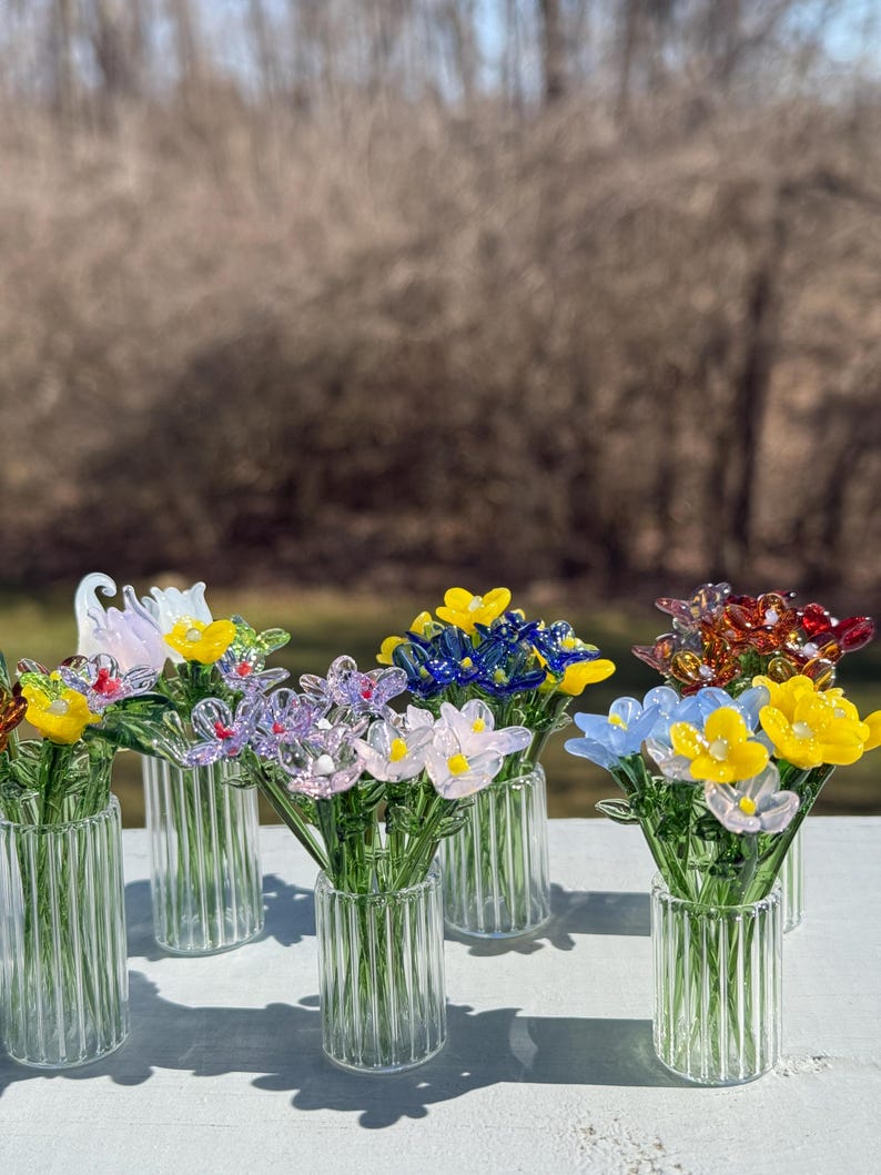 K&ouml;nnte beinhalten: Eine Sammlung bunter Glasblumenstr&auml;u&szlig;e in klaren Glasvasen mit gerippter Textur. Die Blumen sind in verschiedenen Farben angeordnet, darunter Gelb, Rosa, Blau, Lila und Rot.