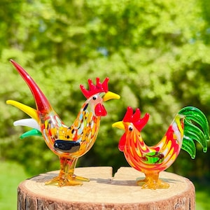 May include: Two colorful glass rooster figurines with red, yellow, and green accents. The roosters are perched on a wooden tree stump against a blurred green background. One rooster has a long, curved tail.