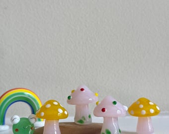 Può includere: Cinque funghi in miniatura di vetro, due gialli con puntini bianchi e tre rosa con puntini verdi e rossi. Una rana di vetro verde e un arcobaleno sono sullo sfondo.