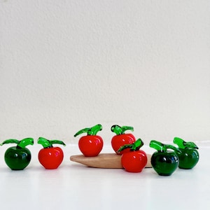 May include: A group of seven glass apples, some red and some green, with green stems and leaves. The apples are arranged on a wooden surface.