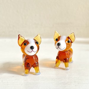 May include: Two small glass figurines of brown and white dogs with black eyes. The dogs are standing on a white surface.