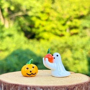 以下が含まれることがあります： ハロウィーンをテーマにしたガラスアートディスプレイ。笑顔のオレンジ色のカボチャと、小さなオレンジ色のカボチャを持った白い幽霊が特徴です。フィギュアは、ぼやけた緑色の背景に対して、木製の表面に置かれています。