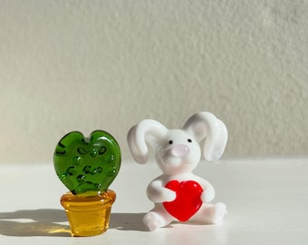 May include: A white glass bunny figurine holding a red heart, next to a green glass heart-shaped plant in a yellow pot.