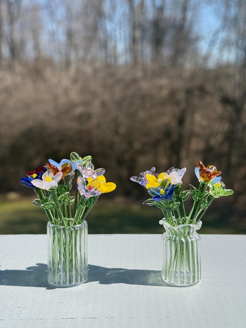 K&ouml;nnte beinhalten: Zwei Glasvasen mit bunten Glasblumenarrangements. Die Vasen sind aus klarem Glas mit einer gerippten Textur. Die Blumen sind in den Farben Blau, Gelb, Rot, Rosa und Gr&uuml;n.