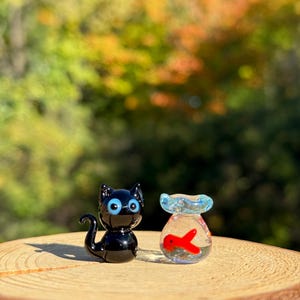 May include: A pair of glass figurines: a black cat with blue eyes and a small fishbowl with a red fish inside. The figurines are displayed on a wooden surface with a blurred background of autumn foliage.