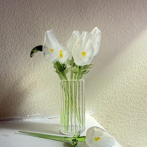 May include: A bouquet of white glass flowers with yellow centres in a clear glass vase with vertical lines. A single white glass flower with a yellow centre lies on a white surface.