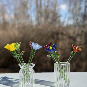 K&ouml;nnte beinhalten: Vier Glasblumen in verschiedenen Farben, gelb, blau, blau und rot, sind in zwei klaren Glasvasen mit geripptem Design angeordnet. Die Vasen stehen auf einer wei&szlig;en Oberfl&auml;che.