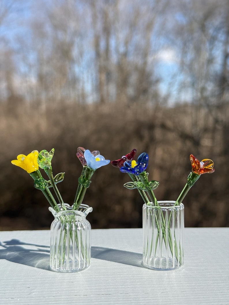 K&ouml;nnte beinhalten: Vier Glasblumen in verschiedenen Farben, gelb, blau, blau und rot, sind in zwei klaren Glasvasen mit geripptem Design angeordnet. Die Vasen stehen auf einer wei&szlig;en Oberfl&auml;che.