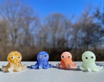 May include: Four colorful glass octopus figurines. The octopus are in shades of yellow, blue, orange, and green. Each octopus has a round head, eight tentacles, and two black eyes. The figurines are displayed on a white surface with a blurred background of trees and a blue sky.