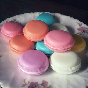 Può includere: Una collezione di colorati macarons nei toni del rosa, arancione, blu e giallo, disposti su un piatto vintage con motivo floreale. I macarons sono impilati, mostrando la loro forma rotonda e la loro farcitura cremosa. Il piatto ha un bordo smerlato e un bordo decorativo rosa.