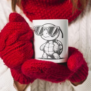 May include: A white ceramic mug with a black and white illustration of a turtle wearing sunglasses. The turtle is standing on its hind legs and has a shell with a pattern. The mug is being held by a person wearing red knitted mittens.
