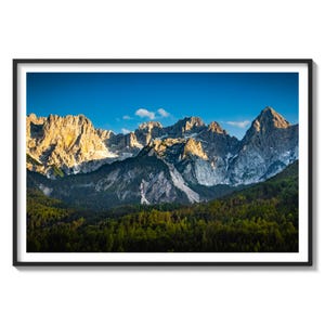 May include: A panoramic view of a mountain range with snow-capped peaks and a lush green forest in the foreground. The sky is a vibrant blue with a few white clouds.