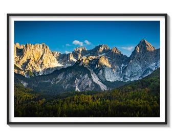 May include: A panoramic view of a mountain range with snow-capped peaks and a lush green forest in the foreground. The sky is a vibrant blue with a few white clouds.