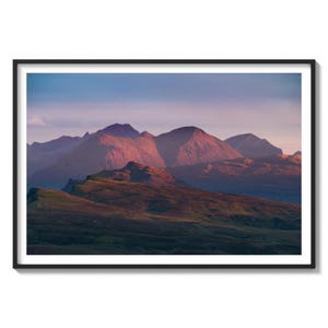 Könnte beinhalten: Eine Panoramaansicht einer Bergkette bei Sonnenuntergang. Die Berge sind in einem tiefen Lila und Rosa gefärbt, mit einem sanften blauen Himmel im Hintergrund. Der Vordergrund ist eine grasbewachsene Wiese.