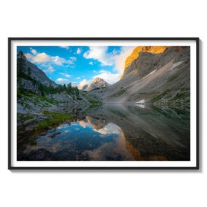 May include: A mountain lake reflecting the sky and surrounding peaks. The water is still and clear, with a bright blue sky and fluffy white clouds reflected in the surface. The mountains are rocky and rugged, with some snow still visible on the peaks. The scene is bathed in the warm light of the setting sun.