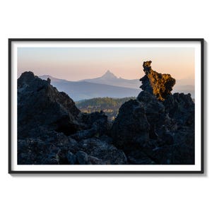 May include: A panoramic view of a mountain range with a rocky foreground. The mountain range is in the distance and appears hazy, while the foreground is made up of dark, jagged rocks. The sky is a soft pink and orange, suggesting a sunrise or sunset.