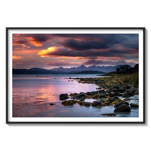 May include: A scenic landscape photograph of a rocky shoreline with a calm body of water in the foreground. The water reflects the colorful sky, which is filled with dramatic clouds at sunset. In the distance, a mountain range is visible.