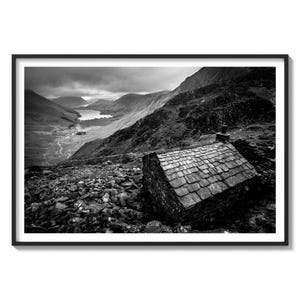 May include: A black and white photograph of a small stone building with a slate roof, perched on a rocky hillside overlooking a valley with a lake in the distance.