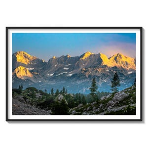 May include: A panoramic view of a mountain range at sunrise, with the peaks illuminated in a warm golden light. The foreground features a lush green forest with trees and rocks.