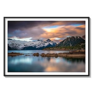 May include: A scenic landscape photograph featuring a mountain range with snow-capped peaks reflected in a still lake. The sky is a vibrant mix of orange, pink, and blue with fluffy clouds.