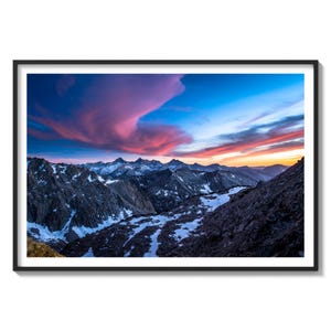 May include: A panoramic view of a mountain range with snow-capped peaks at sunset. The sky is a vibrant mix of pink, orange, and blue with clouds reflecting the colors.